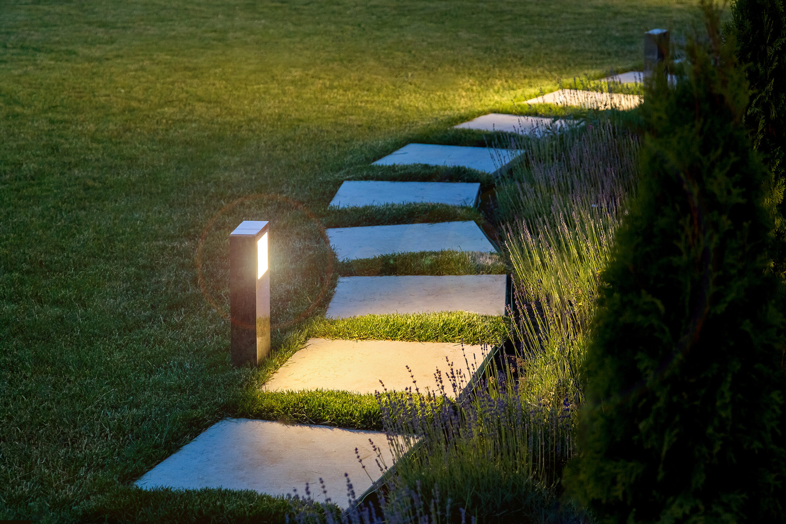 marble path of square tiles illuminated by a lamp glowing with a warm light in a night garden with a flower bed and a lawn, nobody.